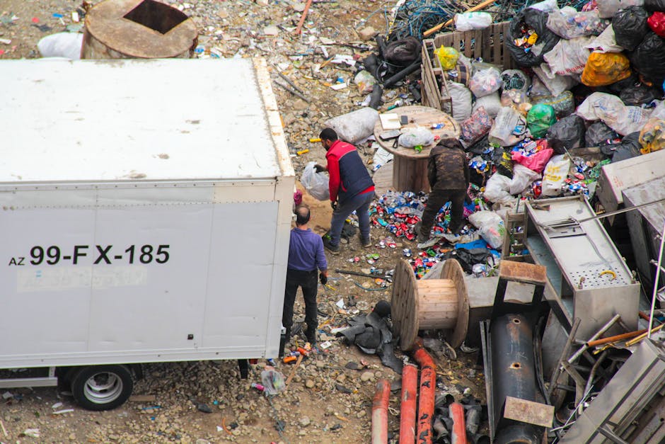 A narrow urban alleyway cluttered with a large, worn black and grey trash bag in the foreground, positioned on a rough asphalt surface with visible dirt and small debris. Behind it, there are piles of