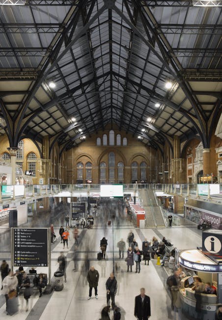 The interior of Liverpool Street station features a high, glass-paneled roof supported by metal beams, allowing natural light to illuminate the spacious, tiled floor. In the foreground, there are seve