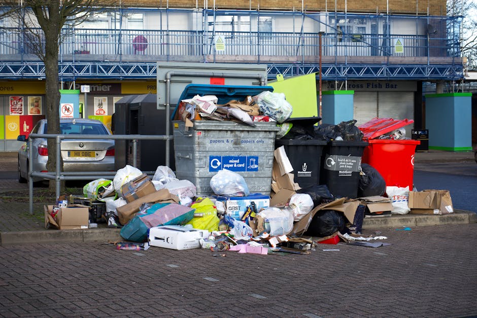 An outdoor scene showing multiple overflowing rubbish bins and scattered waste on the pavement near a parking area. The central large grey bin labeled for mixed paper and card has its lid open, reveal